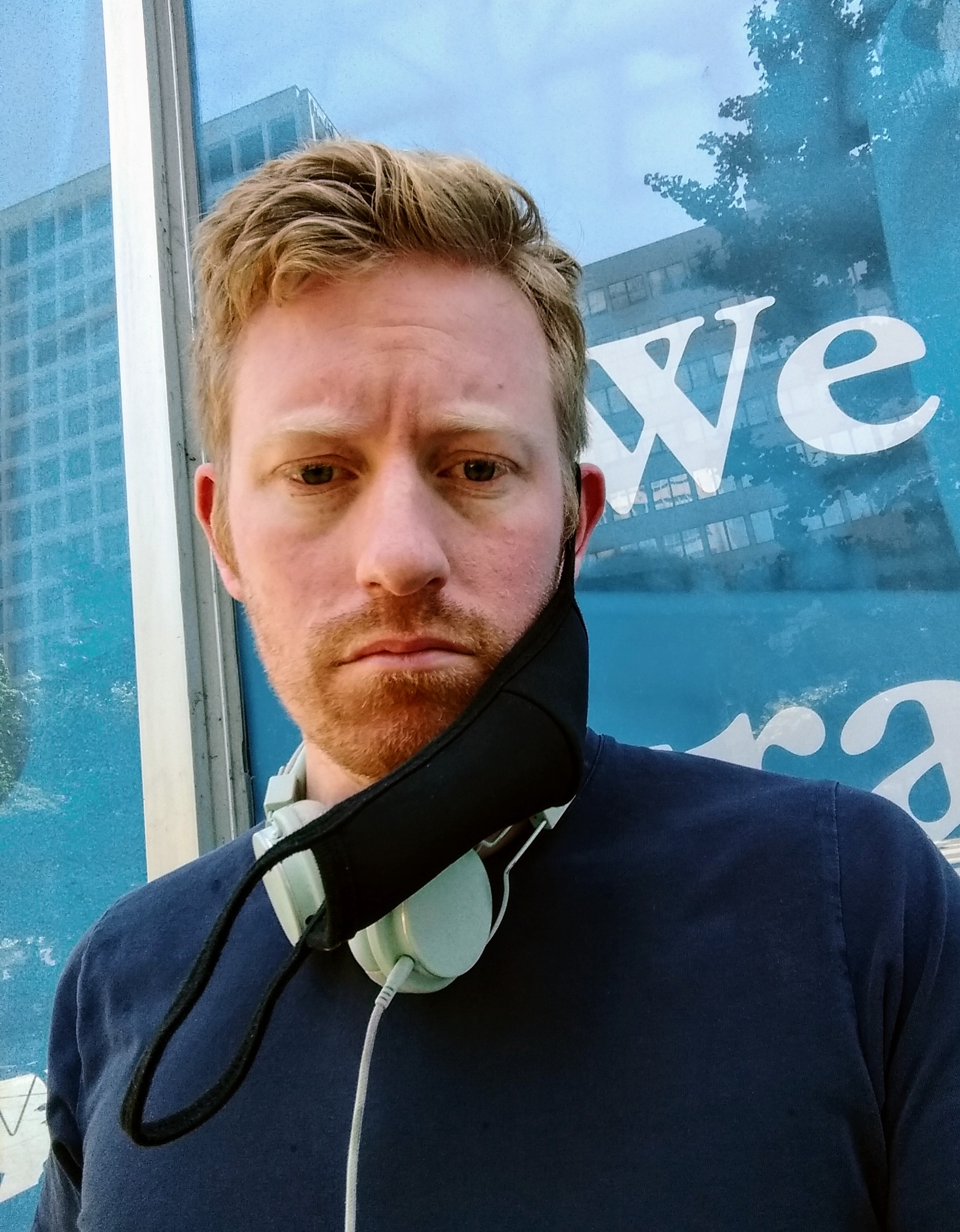 Red-haired young man stoic expression half-wearing a fabric mask.