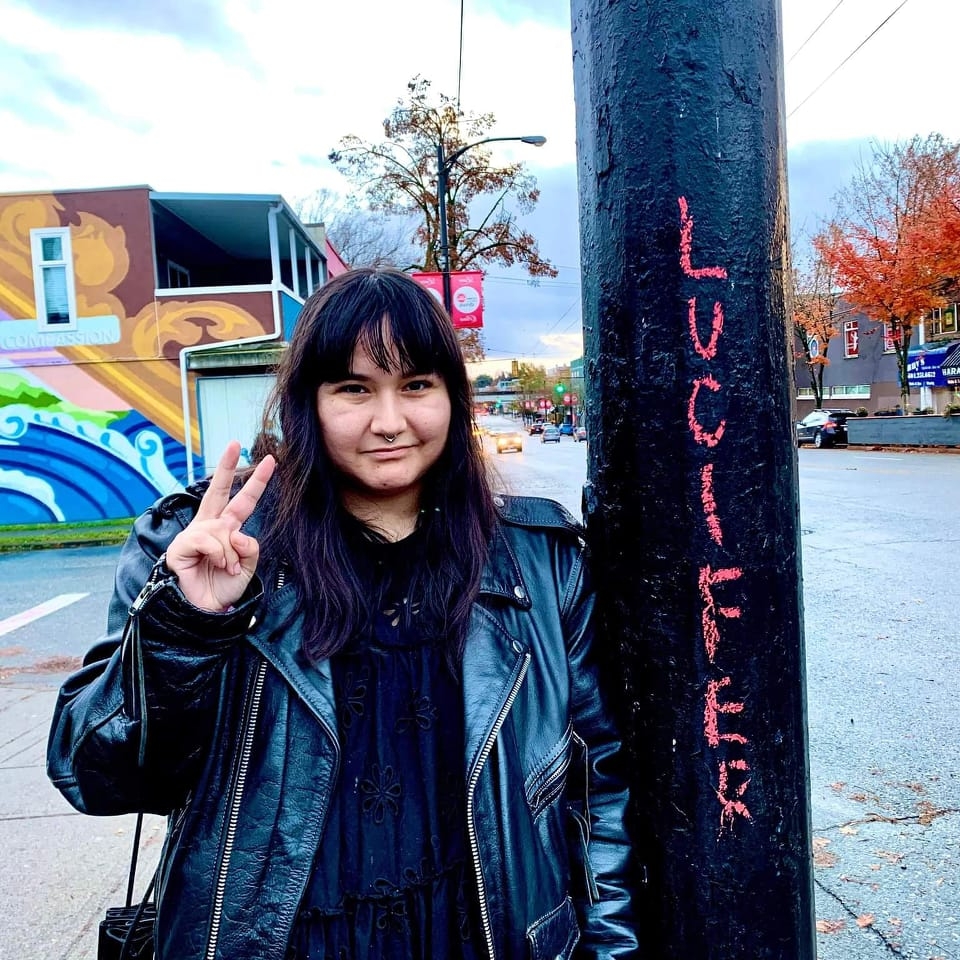 Brandi Bird, giving the peace sign beside a pole that has pink graffiti reading LUCIFER. 