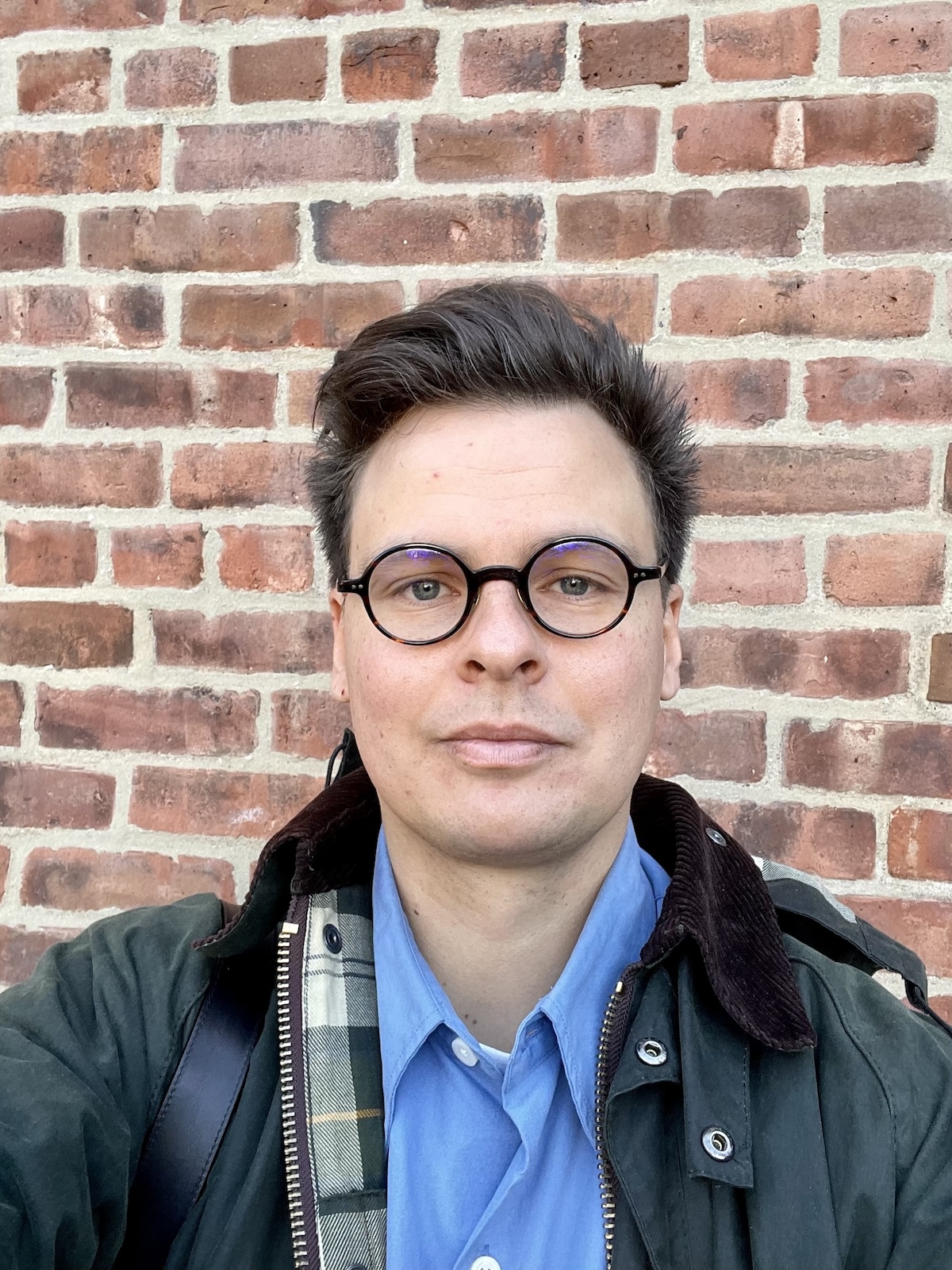 A photograph of a man with brown hair and dark-framed glasses wearing a blue buttondown shirt and a green work coat.