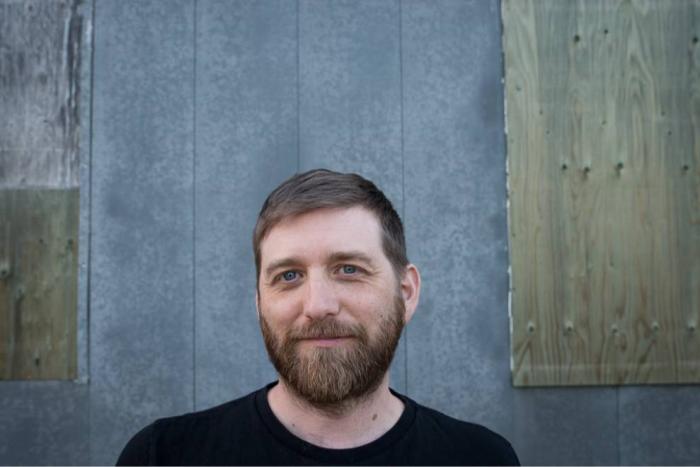 John Samson Fellows stands in front of a gray wall. 
